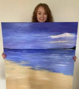 Large lilac acrylic painting of seascape with beach, held by the artist Juliet Firstbrook